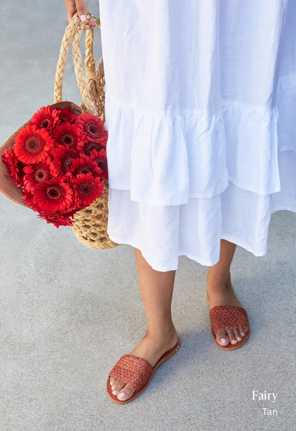 Woman wearing Fairy woven leather slide sandals in tan with white dress, holding flowers.