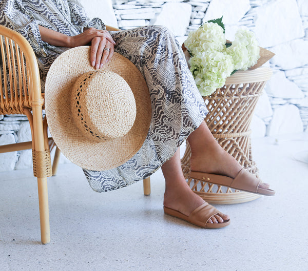 Woman wearing Gallito slide sandals in honey with patterned dress, holding straw hat.