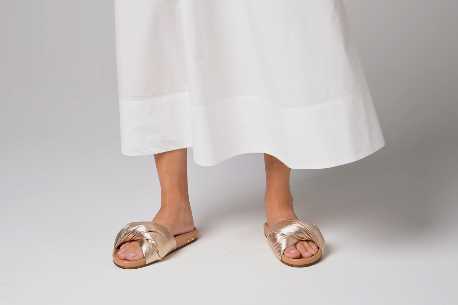Woman walking in Tesia leather slide sandals in gold/beach with white dress
