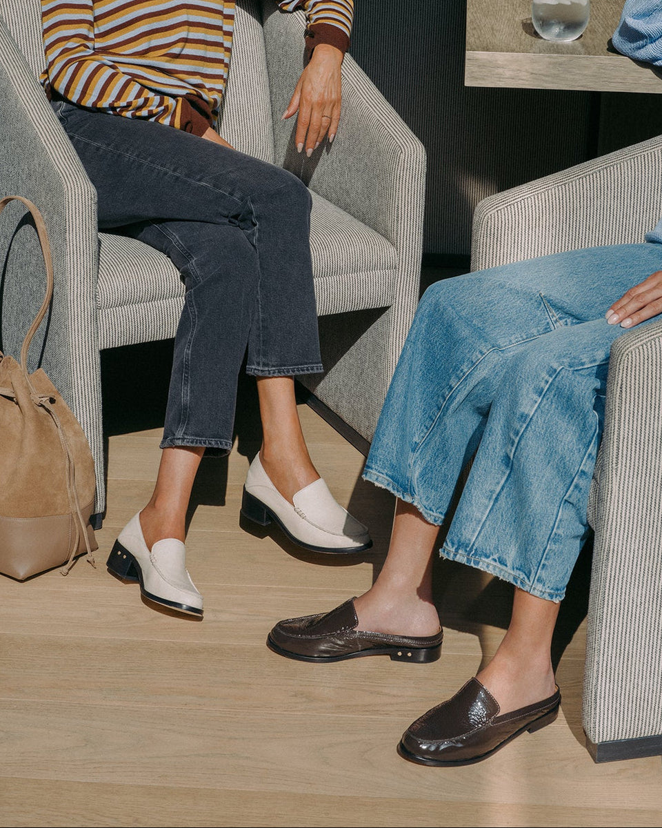 Two models seated in modern gray armchairs in a stylish indoor setting, showcasing different styles of mule loafers. The model on the left wears Owl leather loafer in vanilla with dark cropped jeans and a striped top, while the model on the right wears Owlet backless patent leather loafers with a light blue denim skirt.