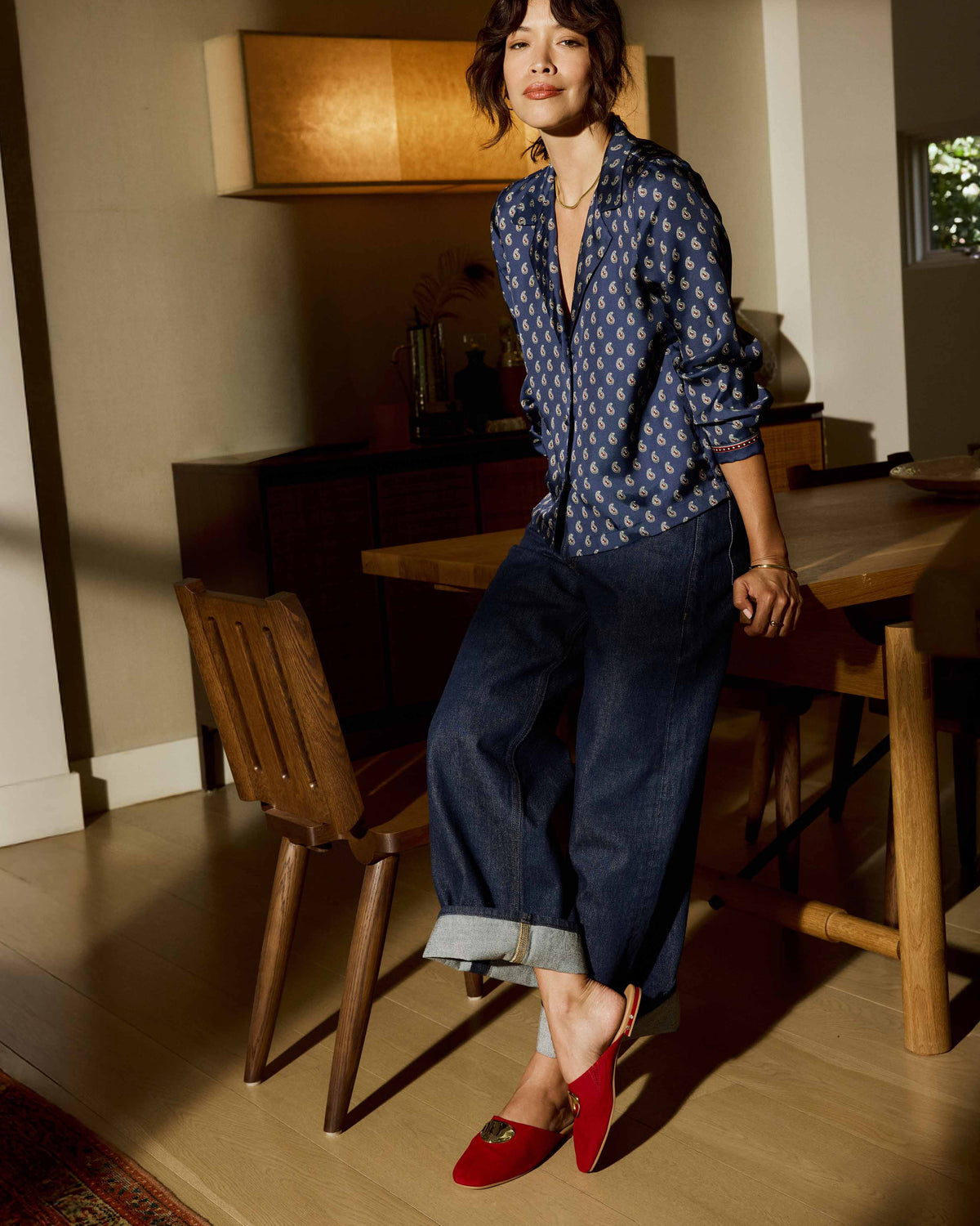 Woman wearing Blackbird Bling suede mule in crimson with attached gold charm with cuffed jeans and paisley blouse