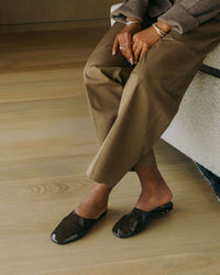 Woman wearing Blackbird patent leather mules in coffee with beige pants and sweater sitting on couch