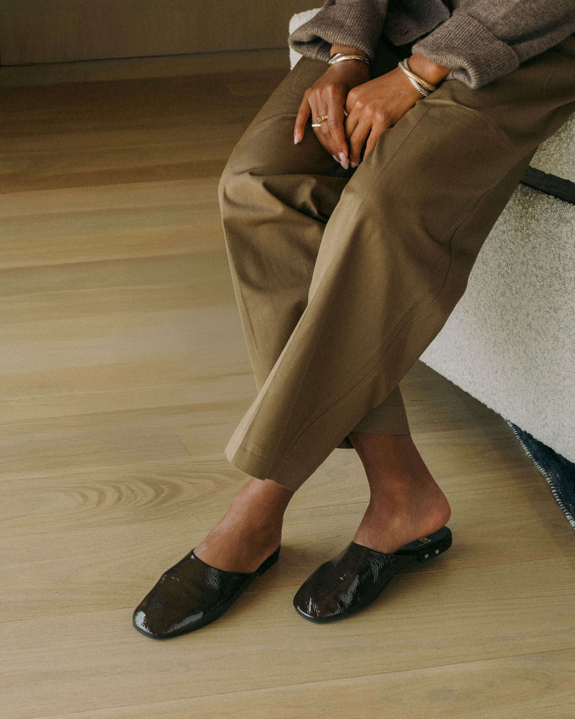 Woman wearing Blackbird patent leather mules in coffee with beige pants and sweater sitting on couch