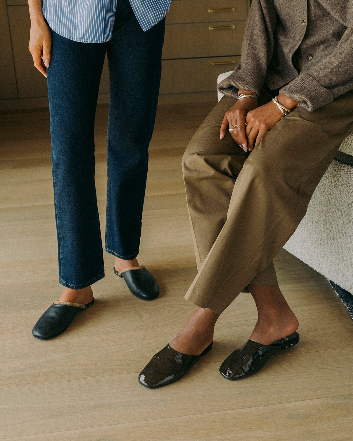 Woman wearing Blackbird patent leather mule in coffee with tan slacks with woman wearing Blackbird Shearling leather mule in black with jeans and striped shirt