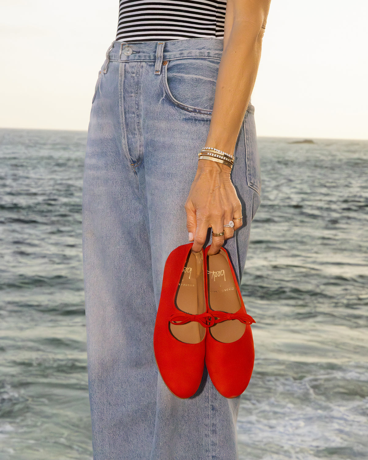 Woman holding Crane Bow velvet mary jane shoes in poppy wearing jeans and striped top by ocean