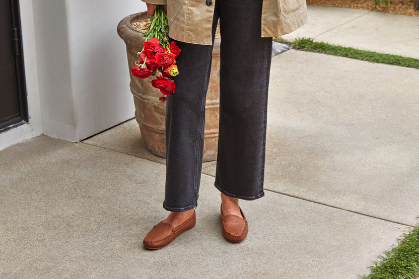 Woman wearing Duck leather loafers in cognac with dark jeans and jacket, holding flowers.