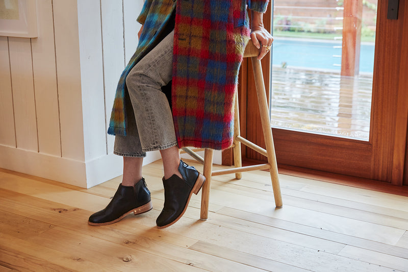 Woman wearing Falcon leather bootie in black with gray jeans and plaid coat, sitting on a stool.