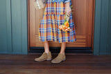 Woman wearing Falcon suede booties in chestnut with plaid dress, holding package and flowers.