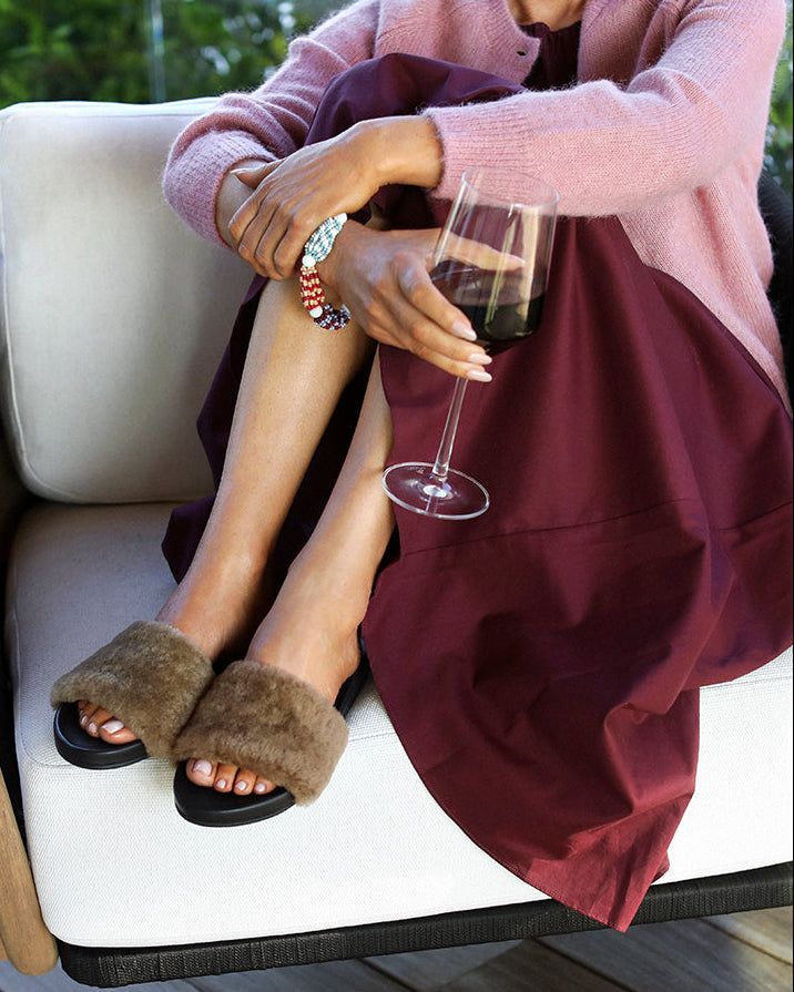 Woman wearing Gallito Más Shearling slide sandals in bronze/black with burgundy dress and pink sweater, sitting in chair, holding glass of wine