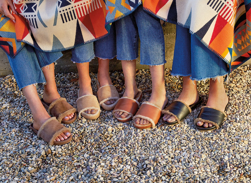 Women wearing Gallito Shearling suede slide sandal in Pecan, Gallito Más Shearling suede slide sandal in almond, Gallito Más Shearling leather slide sandal in cognac, Gallito Shearling leather slide sandal in black with jeans, carrying a blanket.