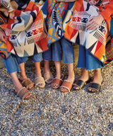 Women wearing Gallito Shearling suede slide sandal in Pecan, Gallito Más Shearling suede slide sandal in almond, Gallito Más Shearling leather slide sandal in cognac, Gallito Shearling leather slide sandal in black with jeans, carrying a blanket.