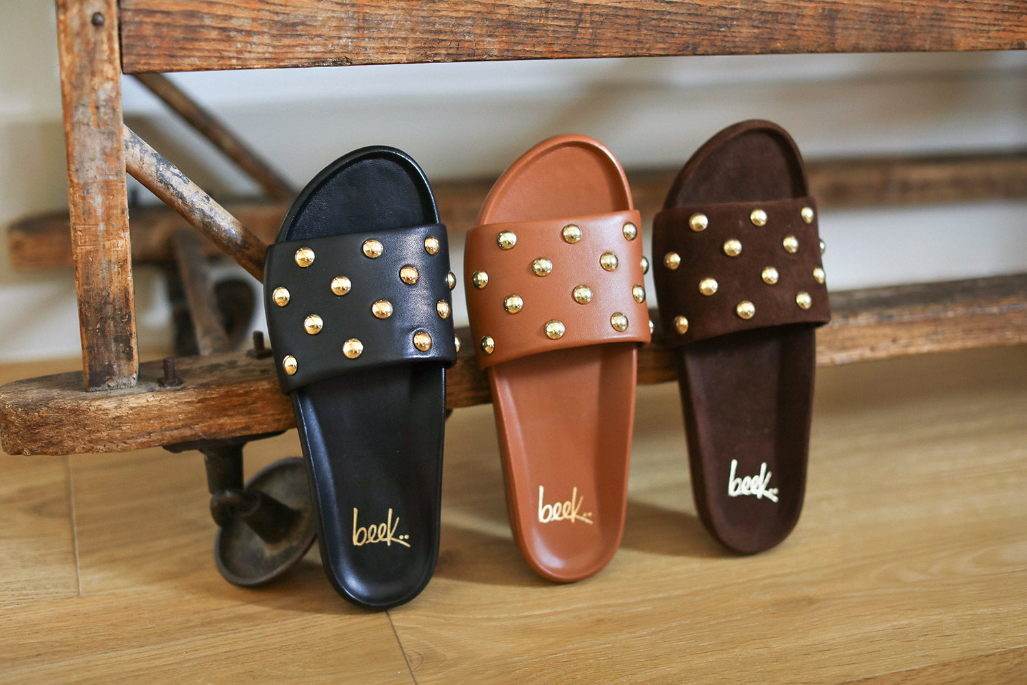 Group shot of Gallito Stud slide sandal in black leather, tan leather, and coffee suede on wood surface.