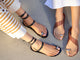 Women wearing jewelry sandal collection: Amethyst leather toe-ring ankle wrap sandal in black with striped pants and Goldfinch leather toe-ring sandal in tan with white dress.
