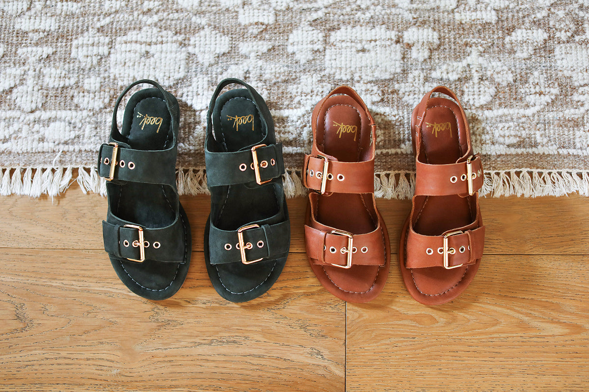 Group shot of Huet buckle sandals in suede ivy and leather cognac shot on a wooden floor with a textured rug in the background.