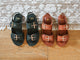 Group shot of Huet buckle sandals in suede ivy and leather cognac shot on a wooden floor with a textured rug in the background.
