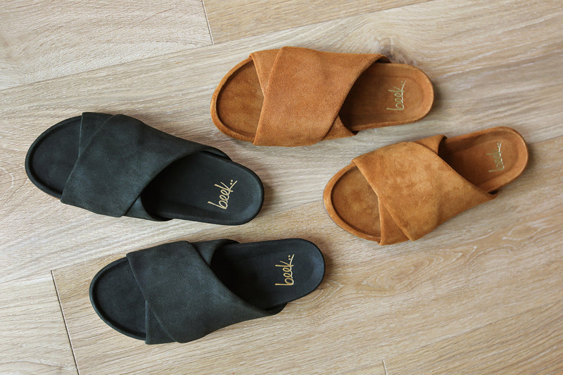 Group shot of Kea suede slide sandals in ivy and caramel on wood surface.