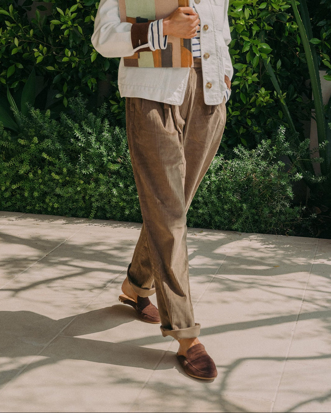 Woman wearing Mallard suede backless loafer mules in coffee with corduroy pants, striped shirt and jacket, holding notebook.