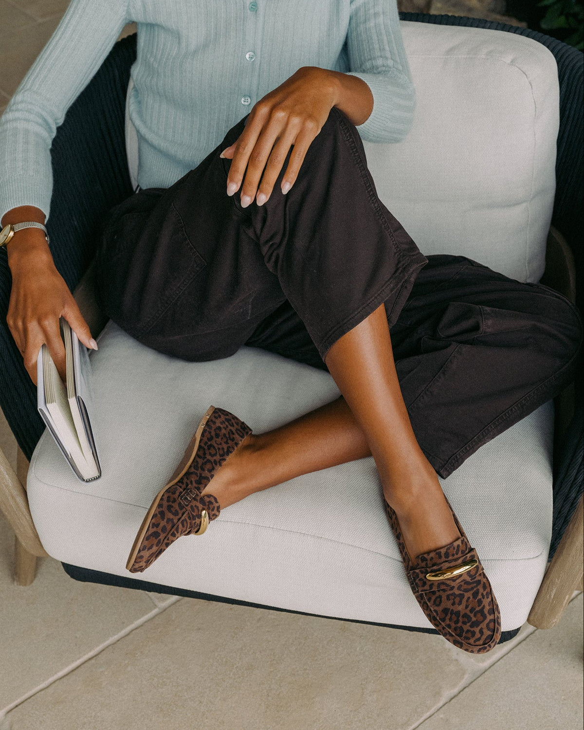 Woman sitting, wearing Moorhen suede loafers in leopard with loafer charm with dark pants and light blue top, holding book.