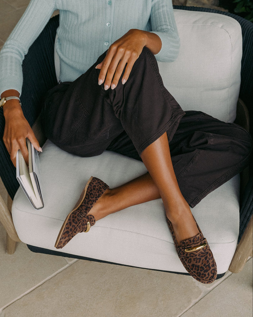 Woman sitting, wearing Moorhen suede loafers in leopard with loafer charm with dark pants and light blue top, holding book.