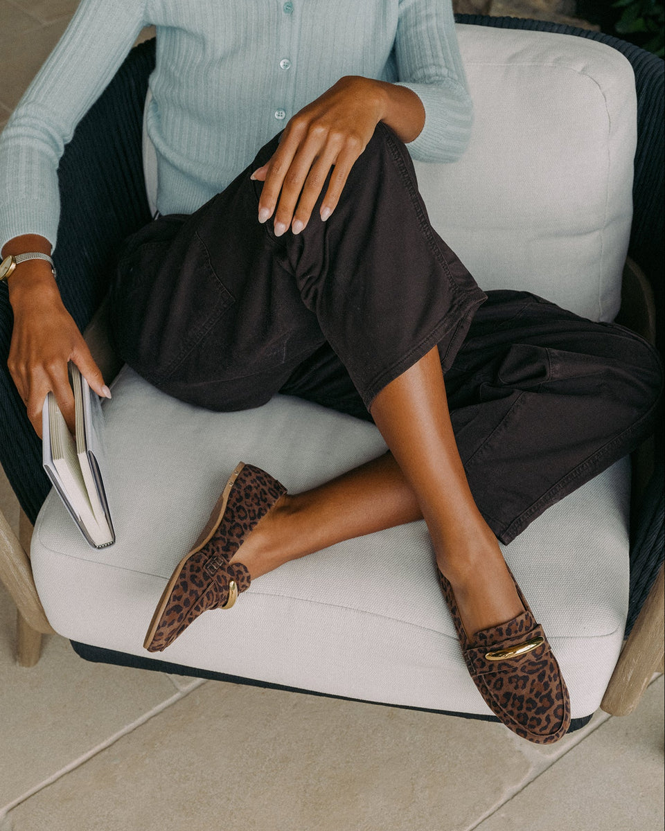 Woman sitting, wearing Moorhen suede loafers in leopard with loafer charm with dark pants and light blue top, holding book.