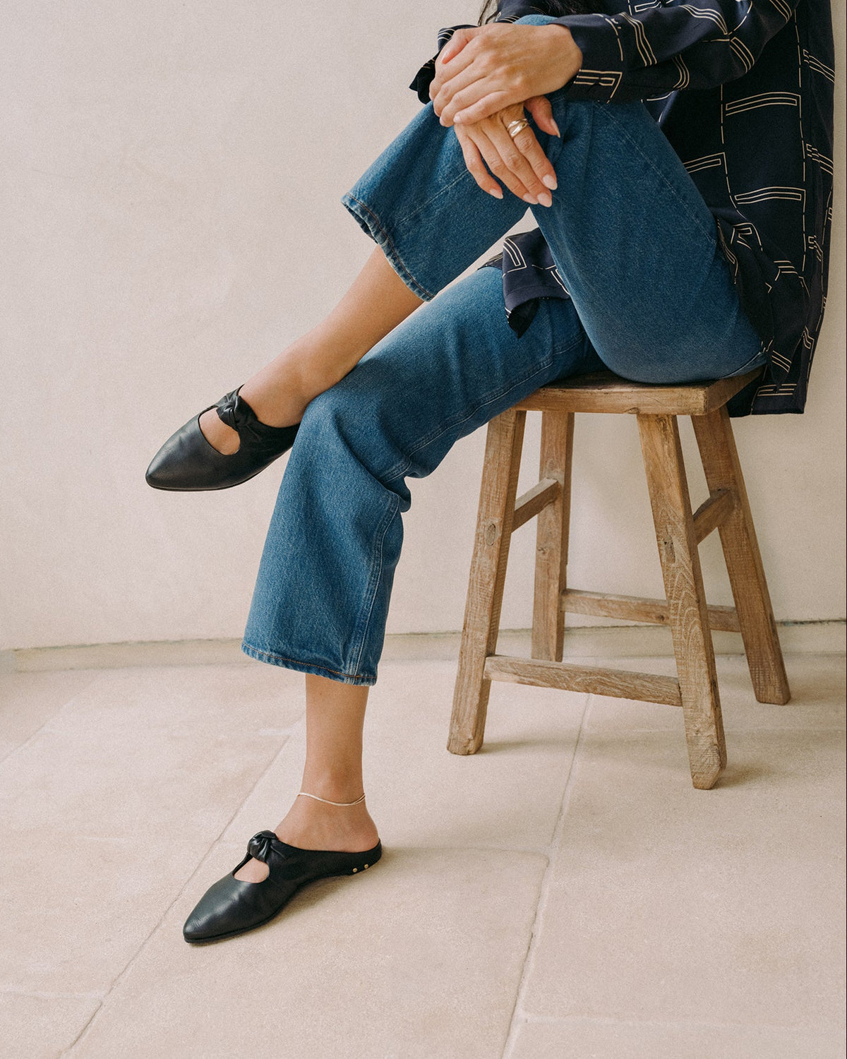 Woman wearing Noddy leather backless ballet mule in black with jeans and patterned top sitting on stool