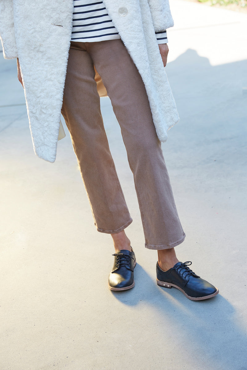 Woman wearing Partridge leather oxford shoes in black with brown pants, striped shirt, and white coat.
