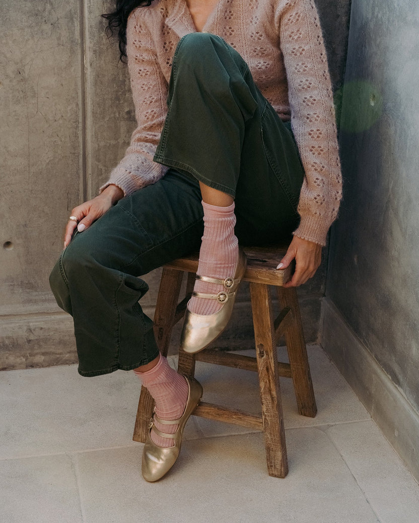 Woman wearing Partridge leather mary jane ballet flats in gold with pants and sweater, sitting on stool.