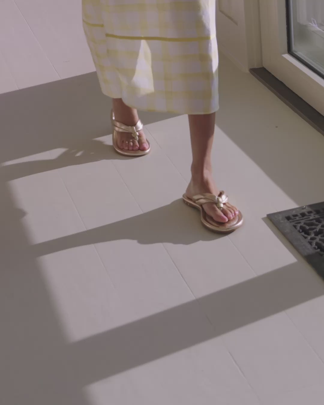 Video of woman wearing Toucan leather flip flop sandals in gold with airy yellow and white checkered dress, walking