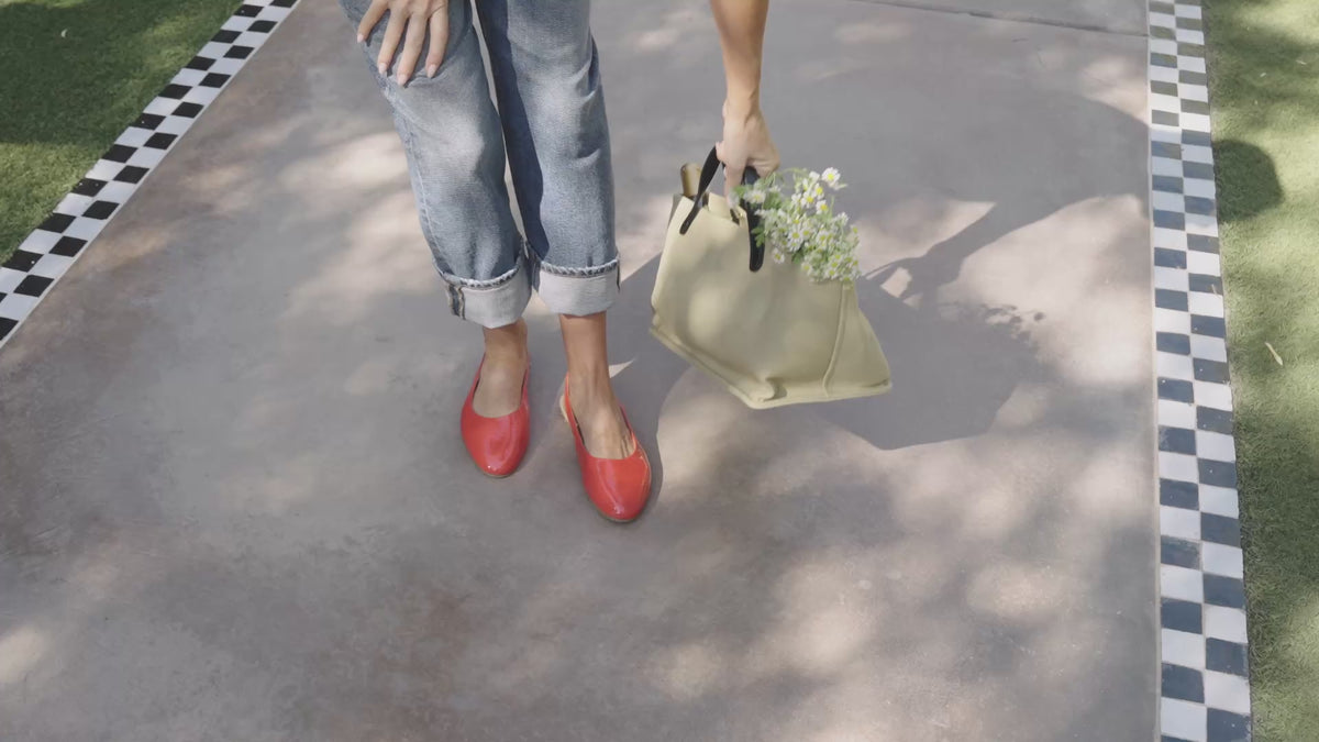 Video of woman wearing Pipit patent leather mules in tomato with jeans, walking and holding a tote bag with flowers
