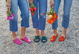 Women wearing Sanderling suede mule in fuchsia, Puffbird suede slide sandal in forest, Moorhen suede loafer in pumpkin and jeans, holding flowers
