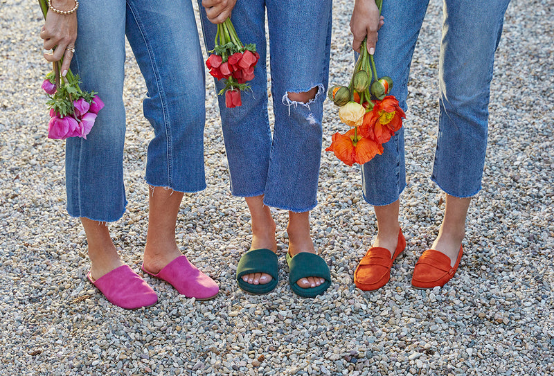 Women wearing Sanderling suede mule in fuchsia, Puffbird suede slide sandal in forest, Moorhen suede loafer in pumpkin and jeans, holding flowers