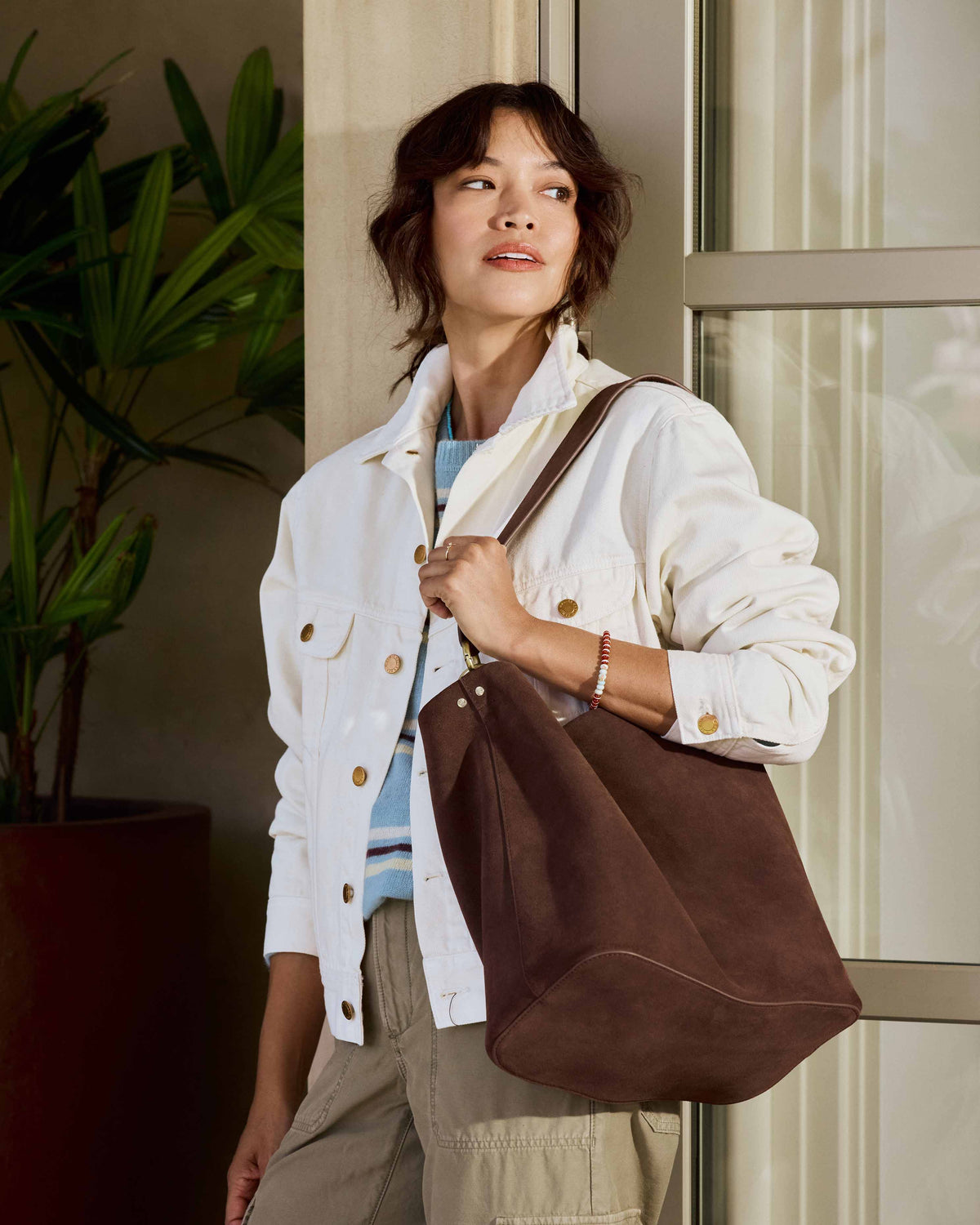 A woman stands in a doorway wearing a white jacket, beige cargo pants, and dark brown loafers, while carrying the Sabine suede bucket bag in coffee