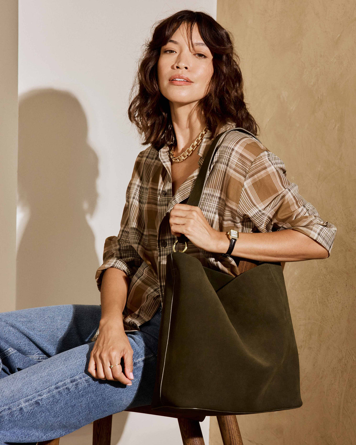 A woman wearing a brown plaid shirt and blue jeans sits on a stool, carrying the Sabine suede bucket bag in olive