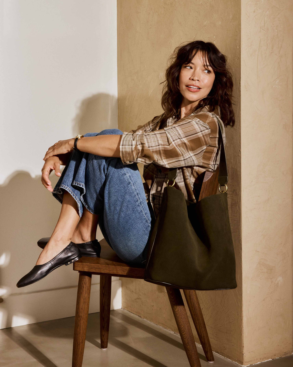 A woman wearing a brown plaid shirt, blue jeans, and Swan leather ballet flats in black sits on a stool, carrying the Sabine suede bucket bag in olive