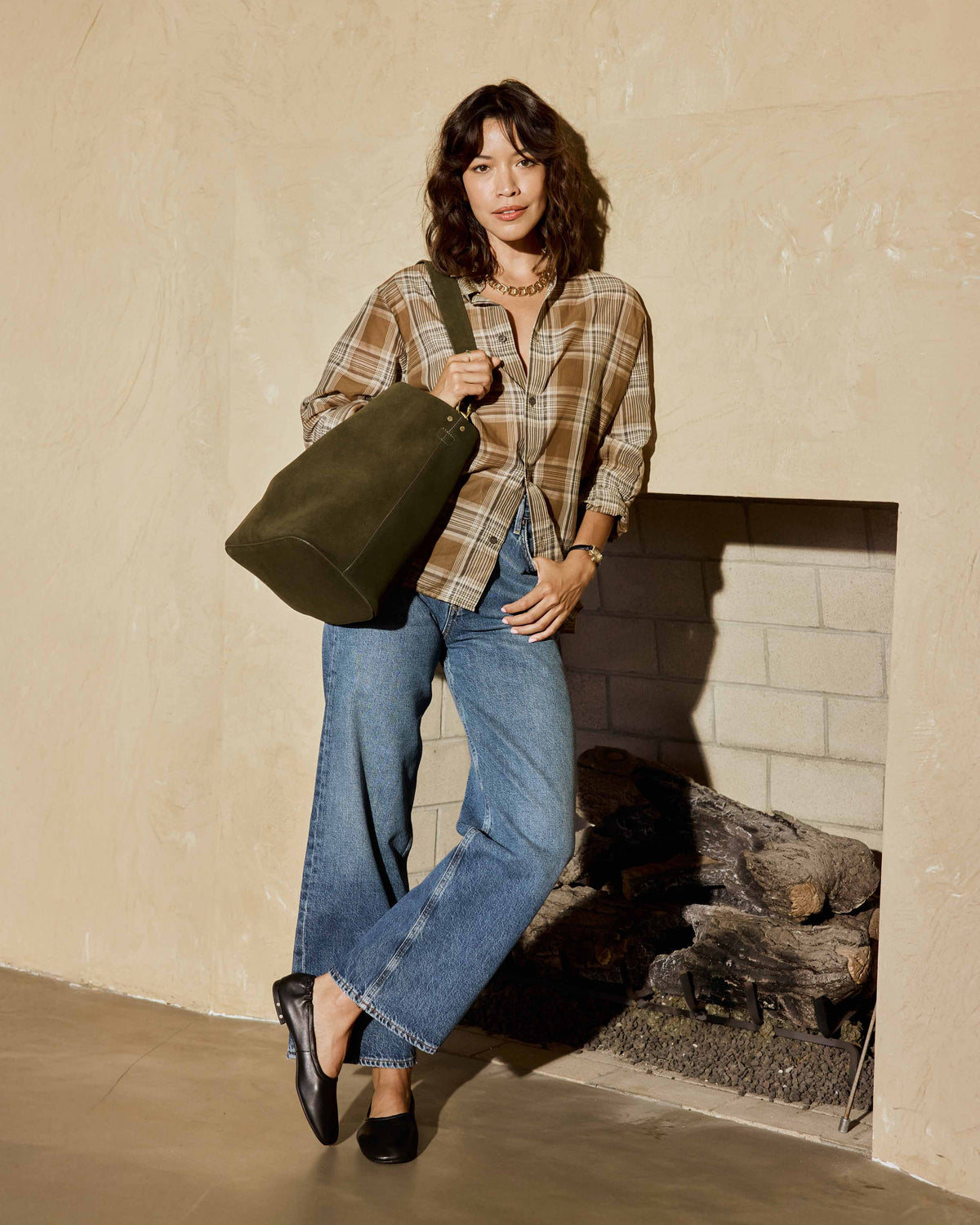 A woman wearing a brown plaid shirt, blue jeans, and Swan leather ballet flats in black stands aginst wall carrying the Sabine suede bucket bag in olive