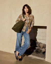 A woman wearing a brown plaid shirt, blue jeans, and Swan leather ballet flats in black stands aginst wall carrying the Sabine suede bucket bag in olive