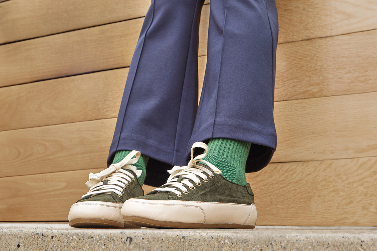 Model wearing Sandpiper suede sneaker shoes in ivy with green socks and blue pants