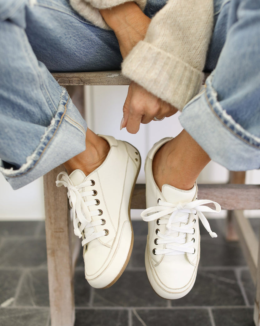 Woman wearing Sandpiper leather sneakers in white with jeans and cream sweater sitting on stool
