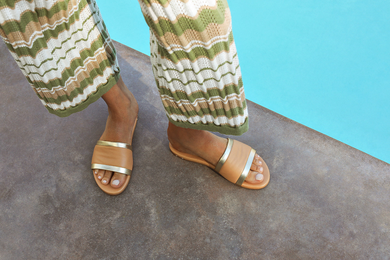 Woman wearing Shorebird leather slide sandal in beach/gold with patterned knit pants