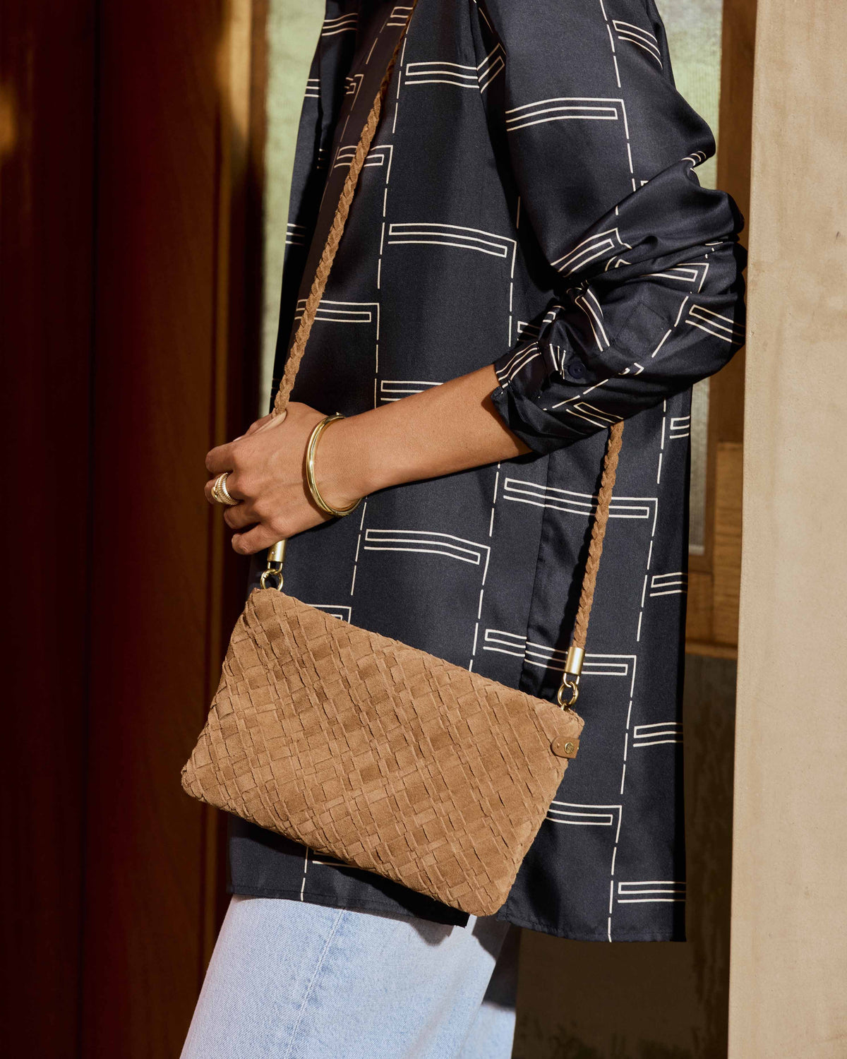 A woman wearing a dark navy patterned shirt and light blue jeans carries the Starling suede woven clutch with strap