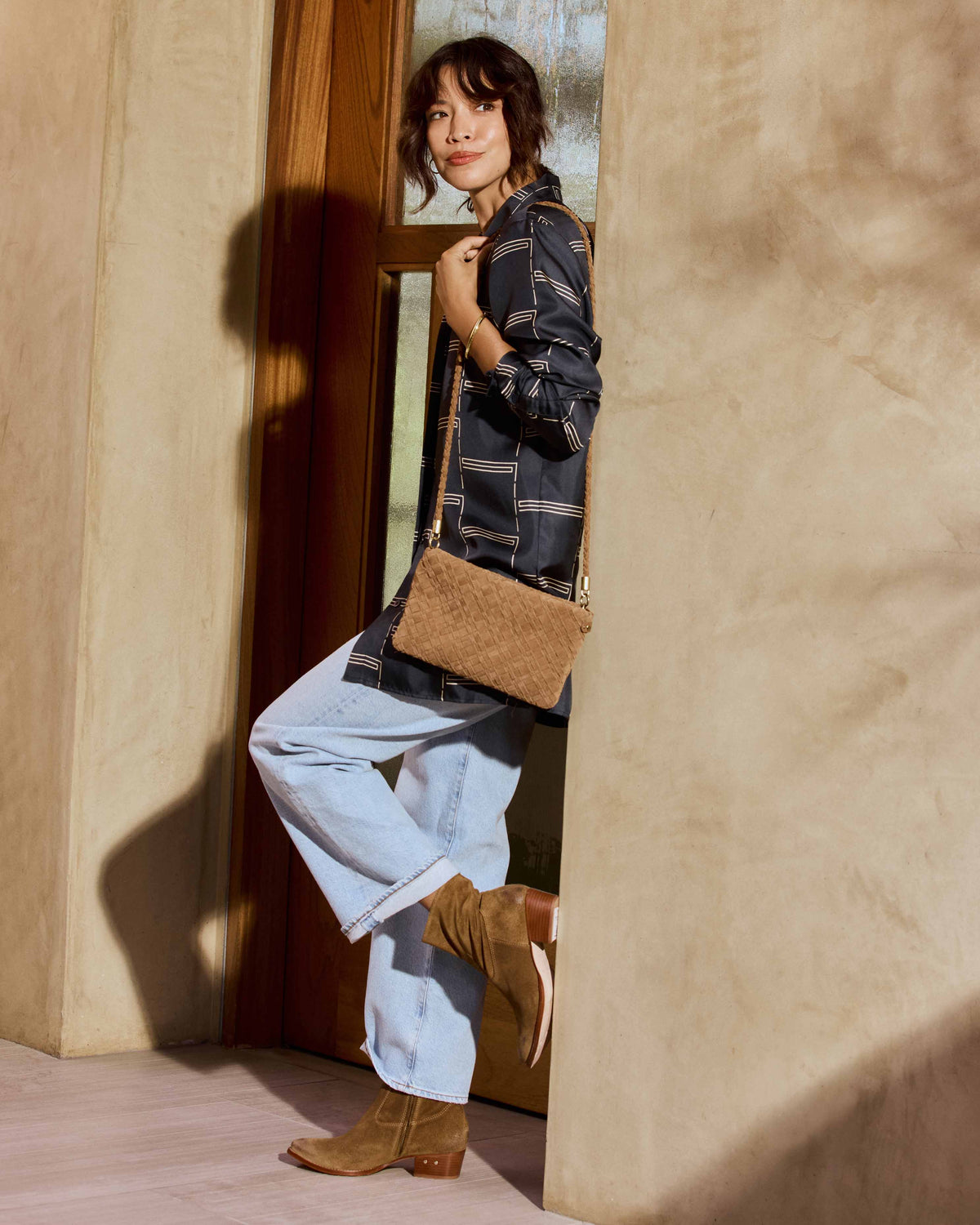 A woman wearing a dark navy patterned shirt, light blue jeans, and Cuervo suede boots in chestnut carries the Starling suede woven clutch with strap in chestnut
