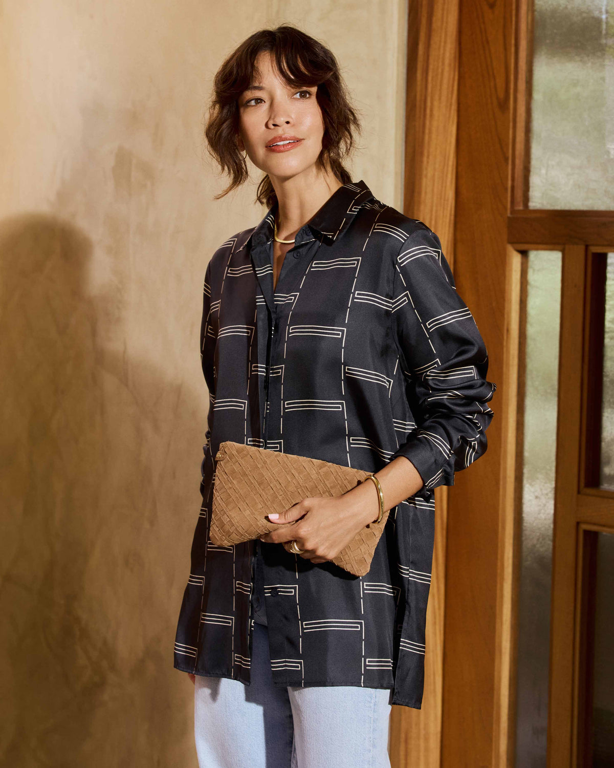 A woman wearing a dark navy patterned shirt, light blue jeans carries the Starling suede woven clutch in chestnut