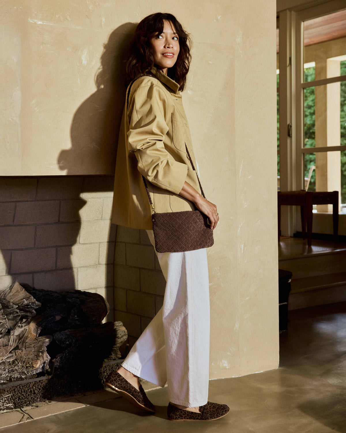 A woman wearing a beige utility jacket, white pants, and Moorhen suede loafers in leopard carries the Starling suede woven clutch with strap in coffee