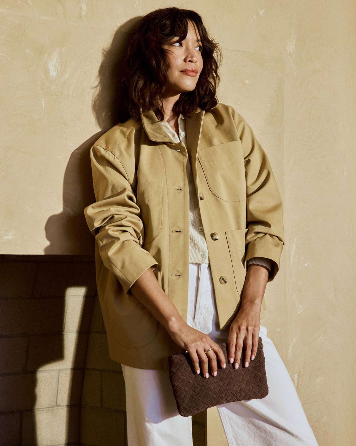 A woman wearing a beige utility jacket, white pants, and Moorhen suede loafers in leopard carries the Starling suede woven clutch  in coffee