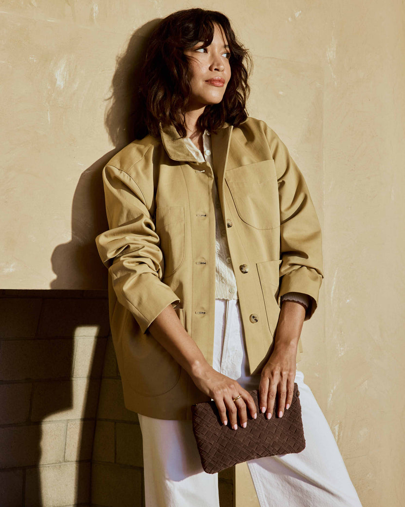 A woman wearing a beige utility jacket, white pants, and Moorhen suede loafers in leopard carries the Starling suede woven clutch  in coffee