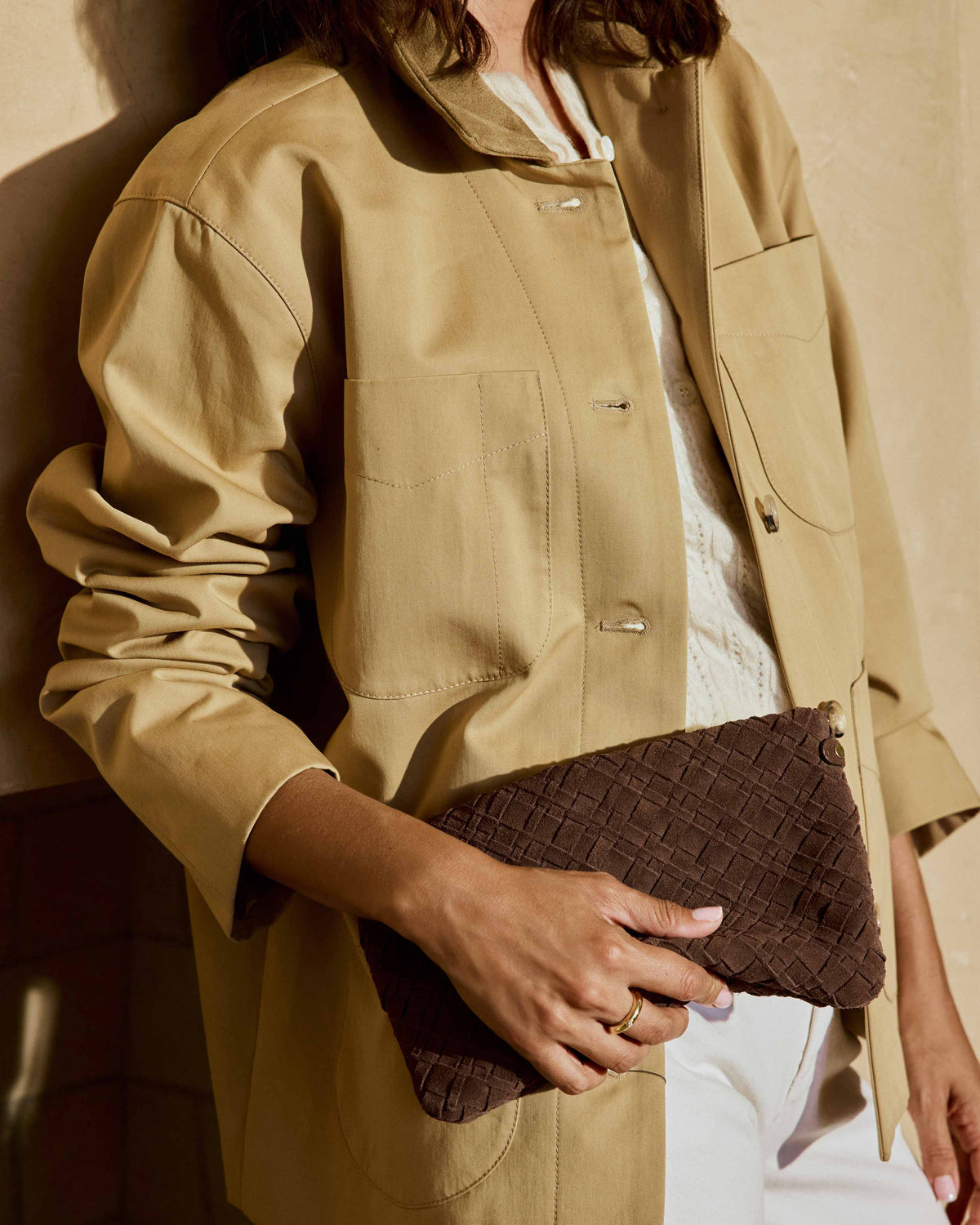 A woman wearing a beige utility jacket, white pants carries the Starling suede woven clutch in coffee