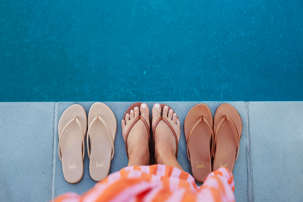 Woman wearing Sunbeam leather flip flop sandal in tan with striped dress next to Sunbeam flip flop sandals in beach and rose gold.