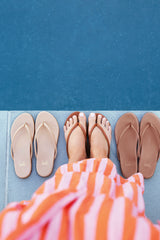 Woman wearing Sunbeam leather flip flop sandal in tan with striped dress next to Sunbeam flip flop sandals in beach and rose gold.