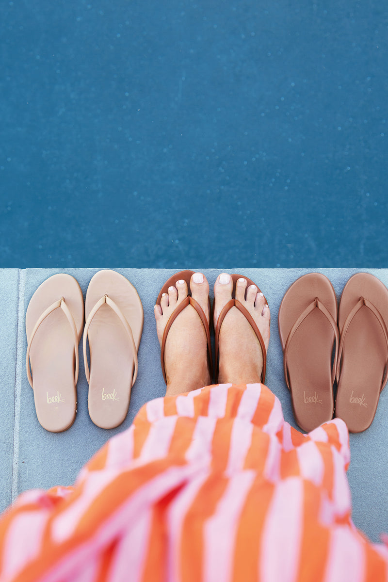 Woman wearing Sunbeam leather flip flop sandal in tan with striped dress next to Sunbeam flip flop sandals in beach and rose gold.