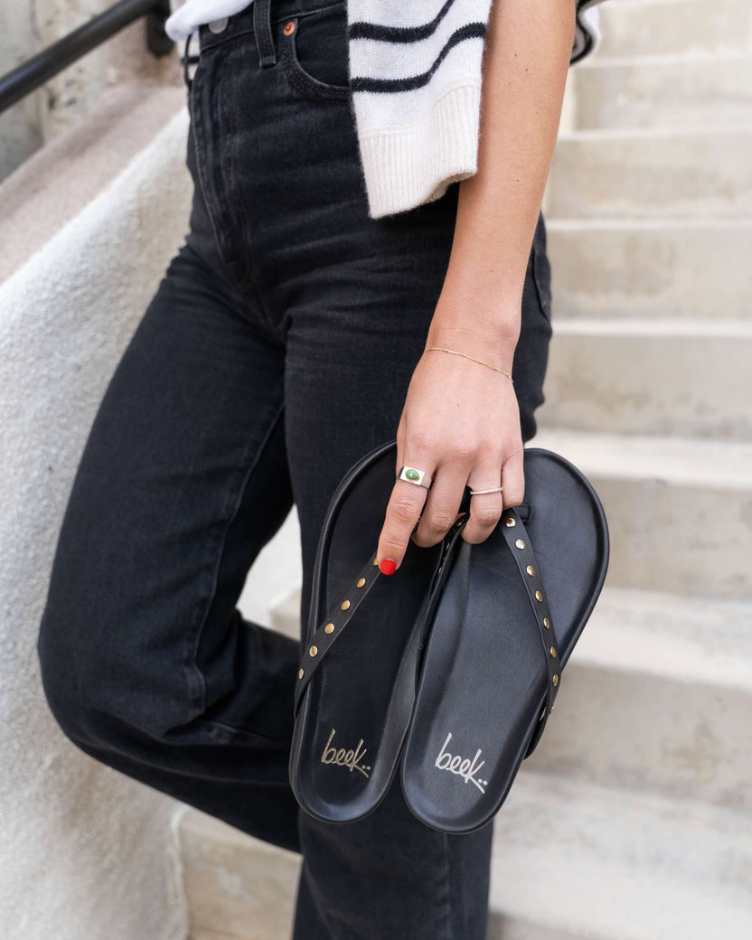 Woman holding Tringa leather flip flop sandal in black with gold studs, wearing black jeans